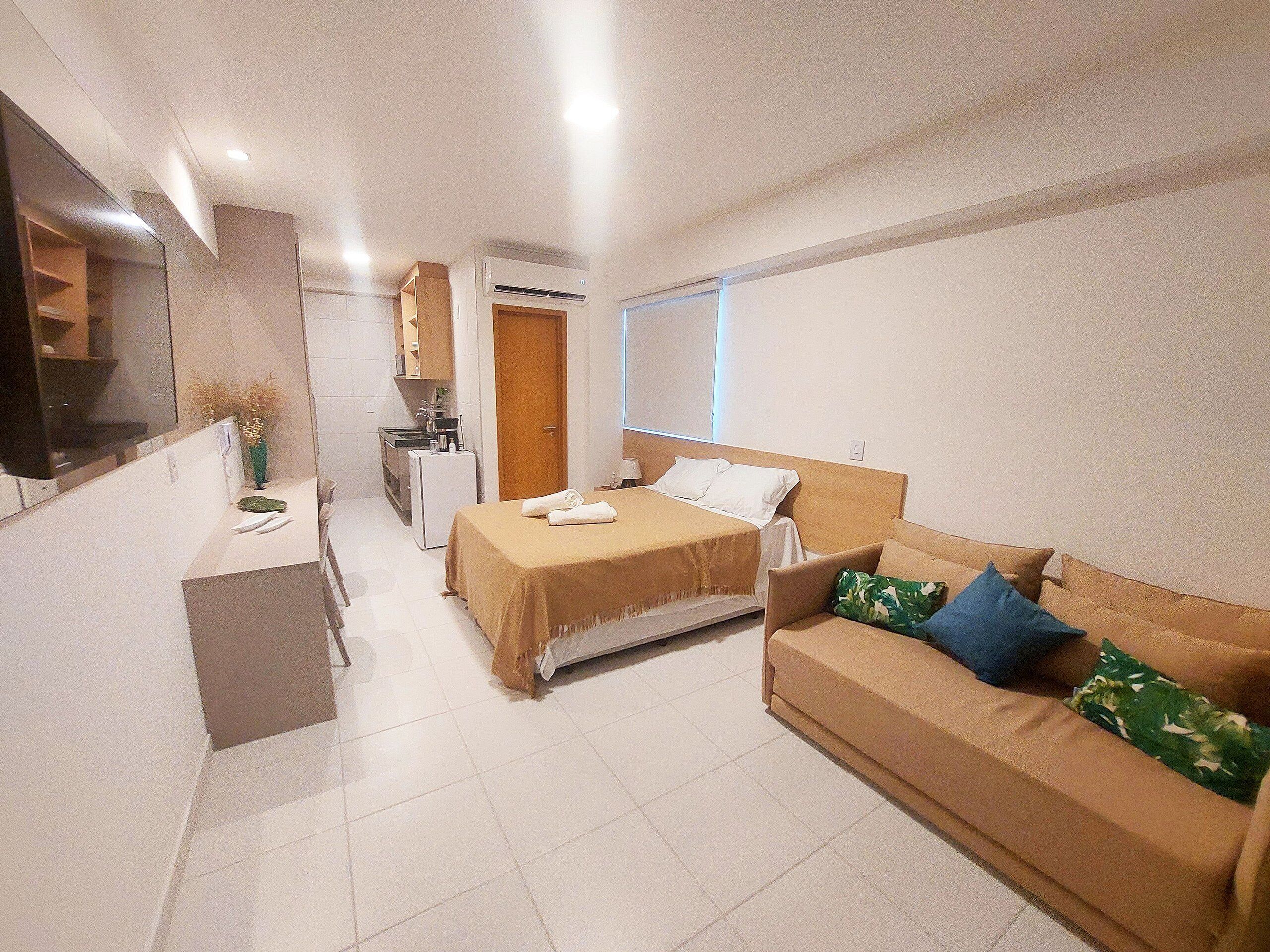 A hotel room with a bed, a sofa, a small kitchenette, a TV, and a window.