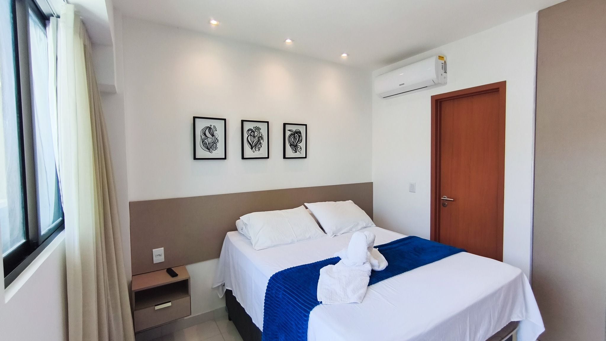 Ein Hotelzimmer mit einem Bett, einem Nachttisch, einer Tür und drei gerahmten Kunstwerken an der Wand.