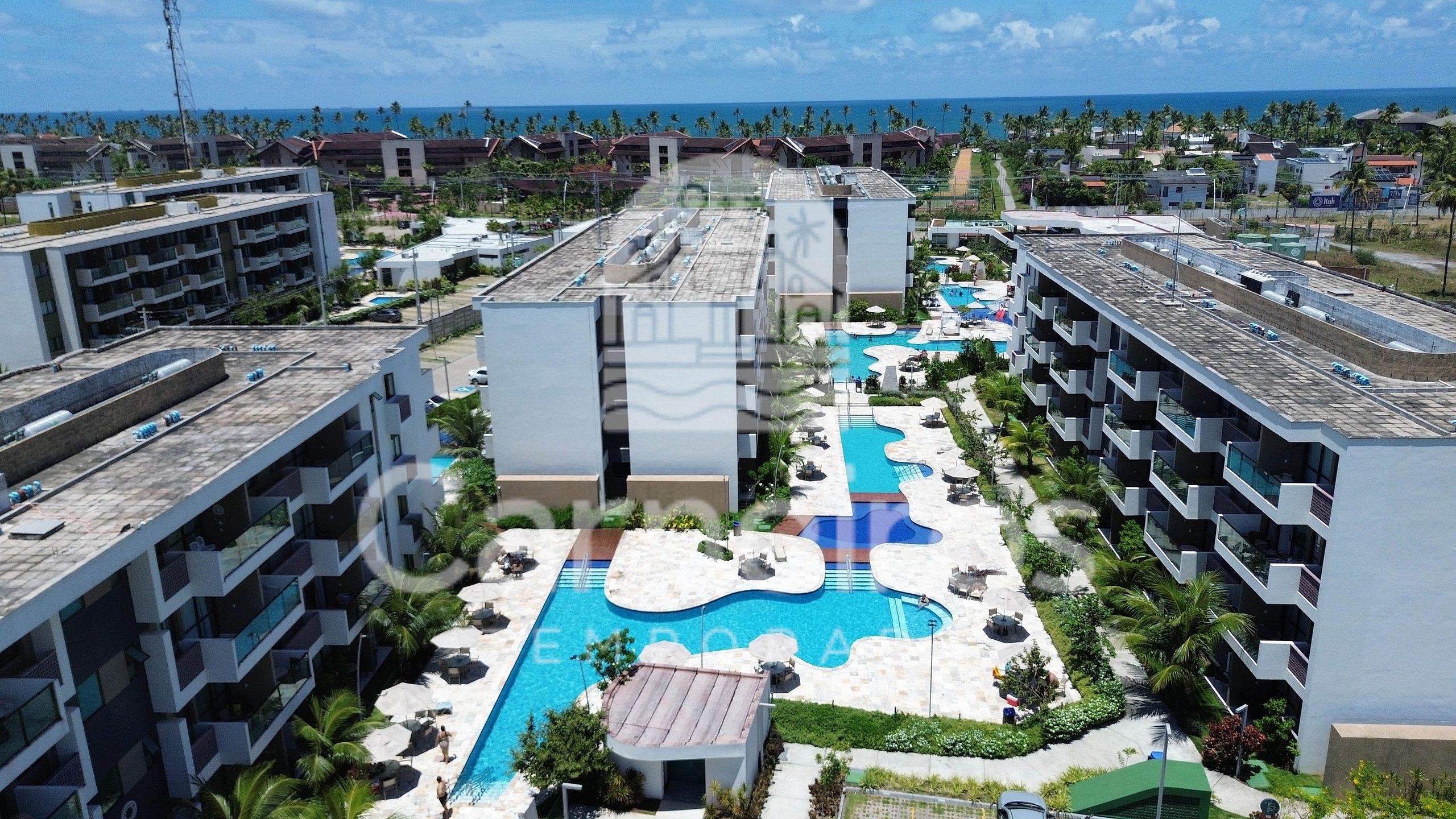 A resort with multiple buildings, a central swimming pool, and a view of the ocean.