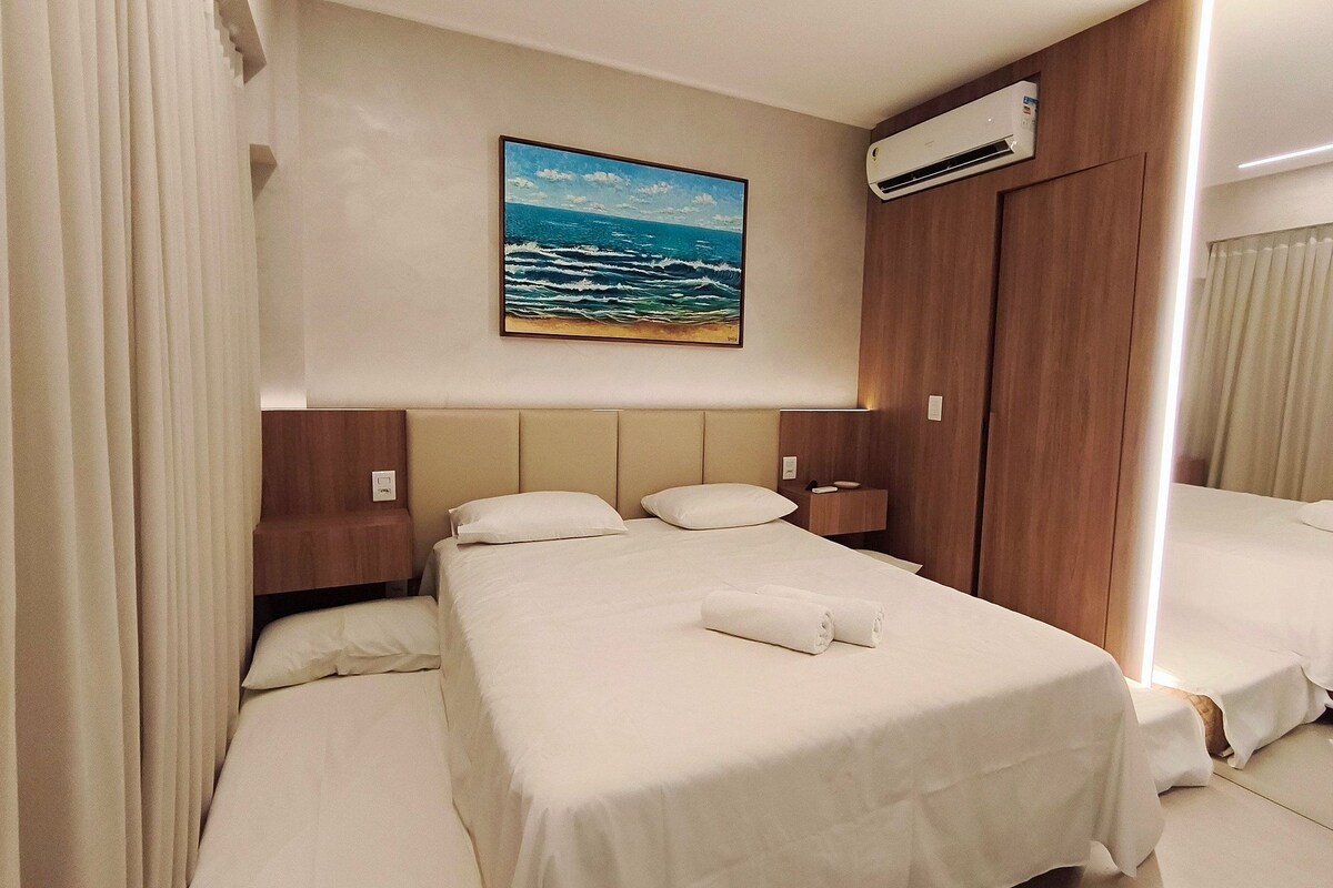 A hotel room with a bed, a nightstand, a painting of the ocean, an air conditioning unit, and a mirror.