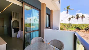 Uma varanda com vista para palmeiras e uma praia, uma mesa redonda com cadeiras, e uma porta de vidro que dá para um quarto com uma cama e uma mesinha.