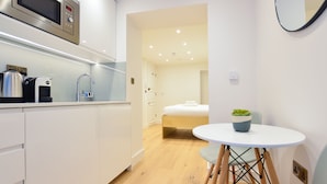 A modern kitchen with a microwave, kettle, and coffee machine. There is a small dining table with a chair and a potted plant. A bed is visible in the background. - Star Street Serviced Apartments by Concept Apartments (London)