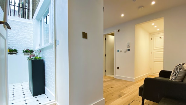A modern interior with a wooden floor, a black sofa, and a small table. There is a door leading to another room and a window with plants on the sill. - Star Street Serviced Apartments by Concept Apartments (London)