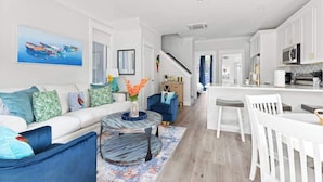 A modern living room with a sofa, coffee table, and chairs, a kitchen with white cabinets and a microwave, and a staircase leading to another level. - Paradise at The Cove Riverwalk Villas (Wilmington)