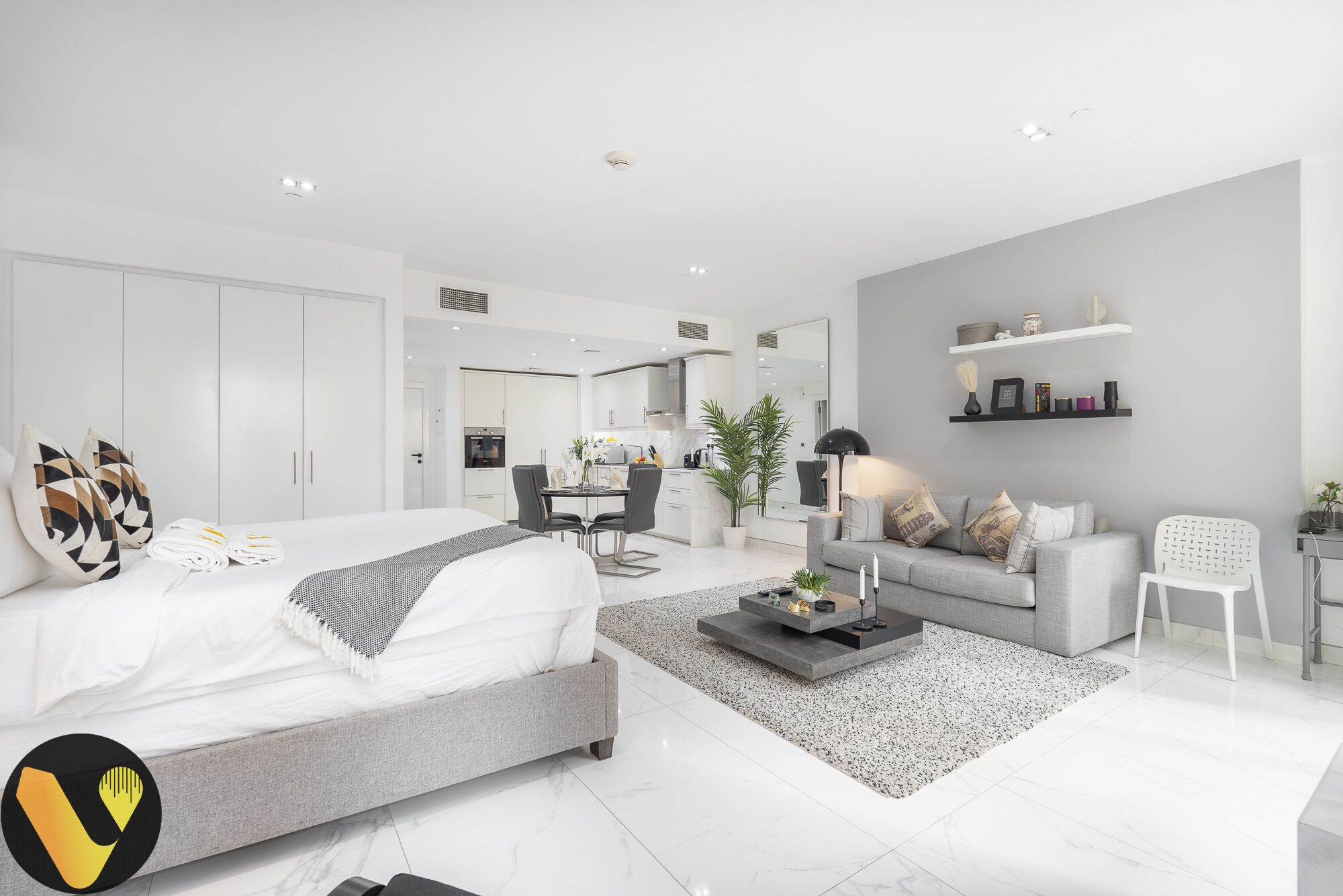 A modern hotel room with a large bed, a dining area, a kitchen, and a living area with a sofa and a coffee table.