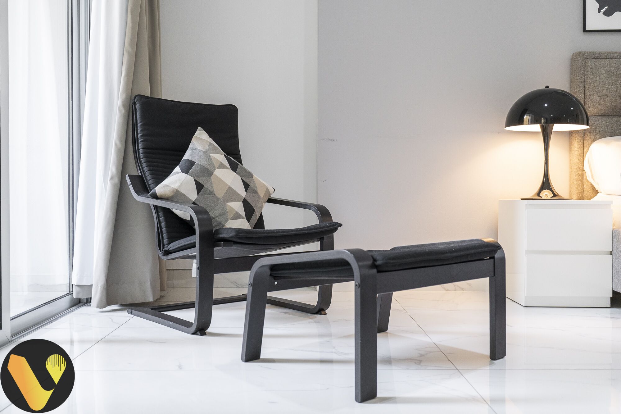 A modern living room with a black leather chair, a patterned cushion, and a black ottoman. A white side table with a lamp is visible.
