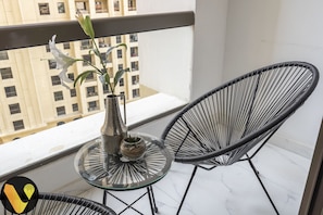 A balcony with a glass table, two black chairs, a metallic vase with a plant, and a view of a building.