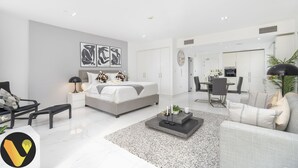Ein modernes, helles Schlafzimmer mit einem großen Bett, Nachttischen und einem Nachtschränkchen. Es gibt einen Essbereich mit Tisch und Stühlen, und im Hintergrund ist eine Küche zu sehen.