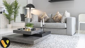 Ein modernes Wohnzimmer mit grauem Sofa, dekorativen Kissen, einem Couchtisch mit Pflanzen, einer Stehlampe und einem Regal mit Büchern und Kerzen.