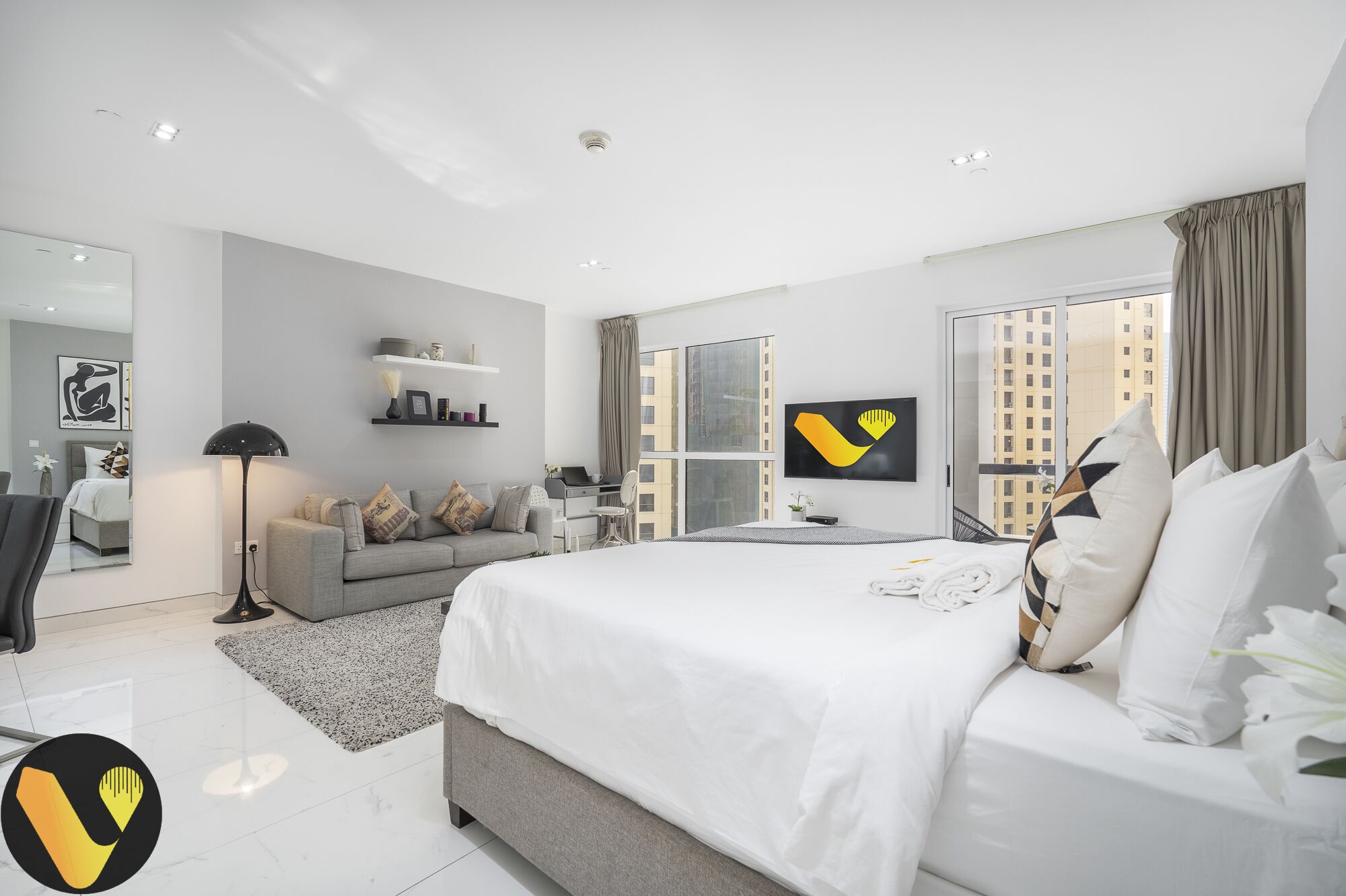 A modern hotel room with a large bed, a desk, and a view of the city through large windows.