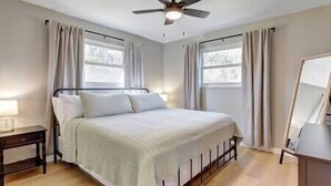 Une chambre à coucher comprenant un lit, une table de chevet, un ventilateur de plafond et deux fenêtres avec des rideaux.