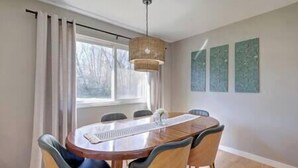Un coin repas comprenant une table ronde en bois, quatre chaises, un luminaire suspendu, une fenêtre avec des rideaux et trois œuvres d’art accrochées au mur.
