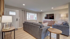Un salon moderne avec un canapé gris, une petite table en bois, un téléviseur fixé au mur et une lampe sur une table d’appoint.