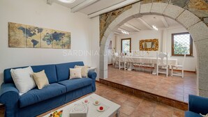 Un salon avec un canapé bleu, une table basse sur laquelle sont disposées des pâtisseries, un coin repas avec une table et des chaises blanches, et une arche en pierre.