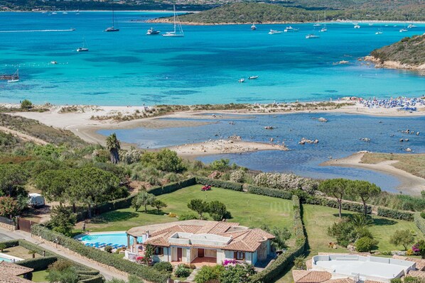 A coastal property with a swimming pool, a large house with a tiled roof, and a view of the sea with boats.