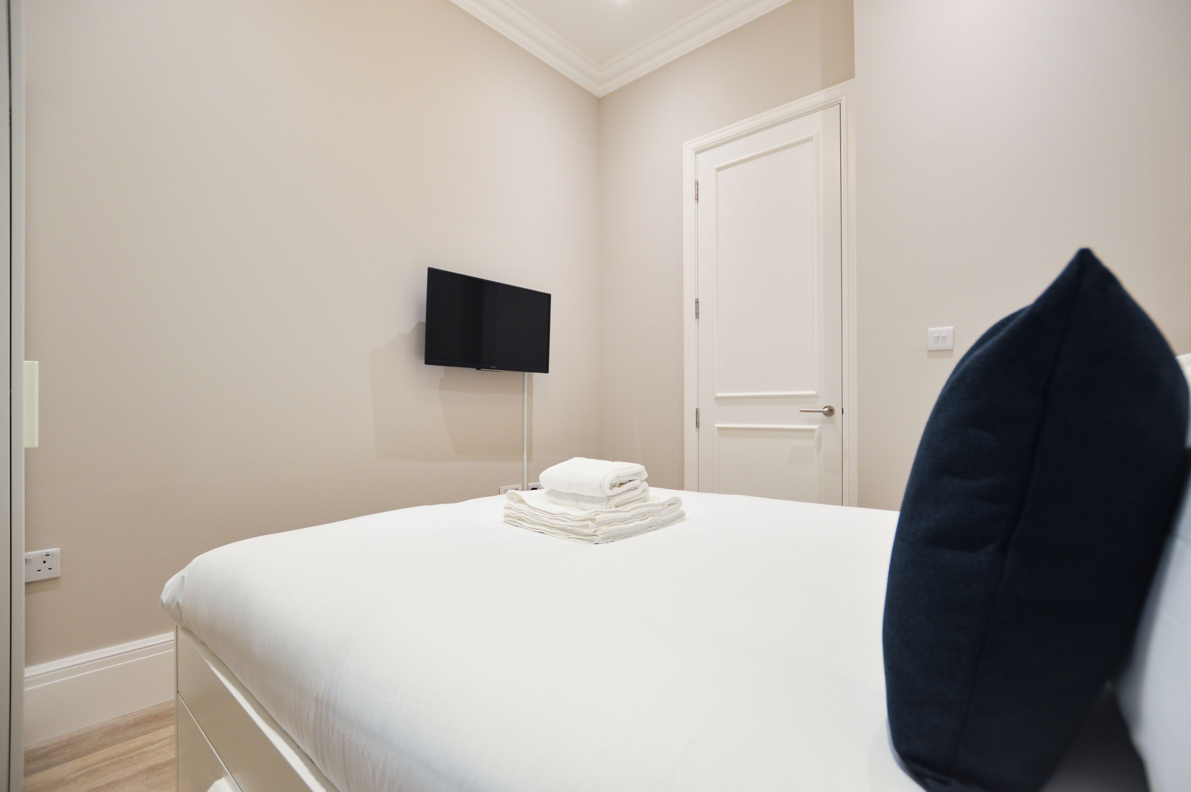 En snyggt bäddad säng med en hopvikt vit handduk, en platt-TV som är monterad på väggen och en stängd dörr.