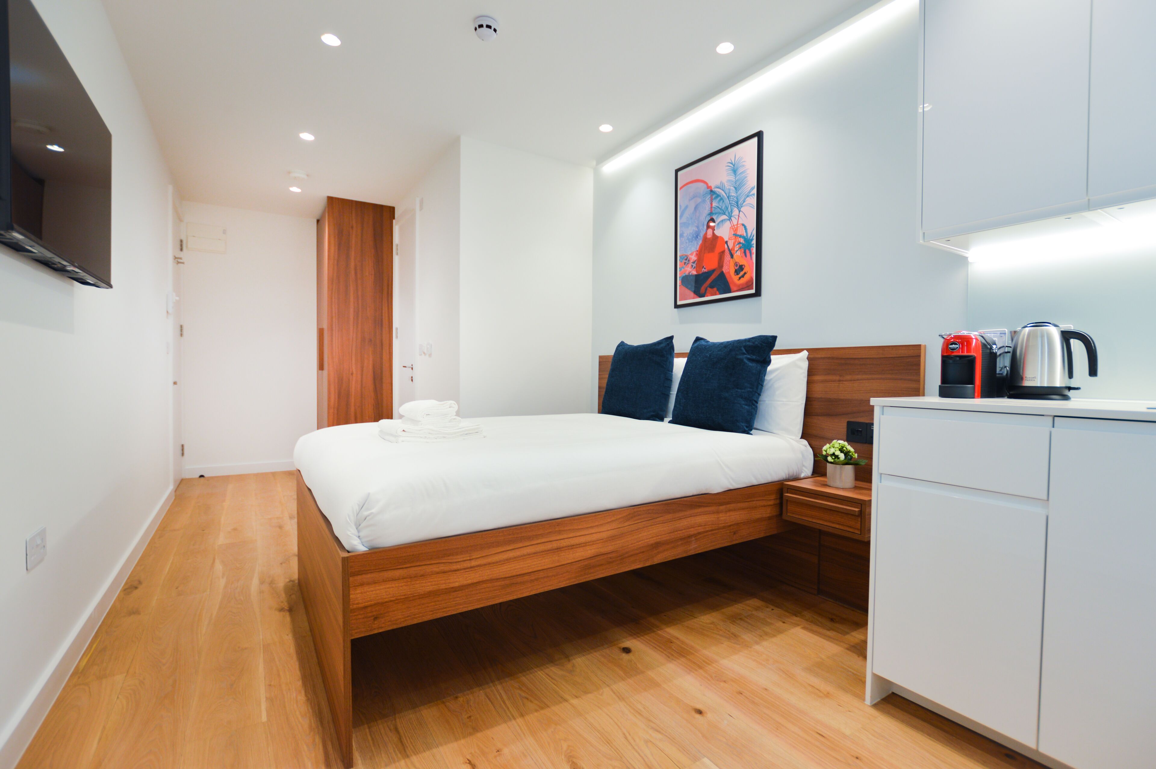 A modern hotel room with a wooden bed, white linens, a nightstand with a kettle, and a painting on the wall.