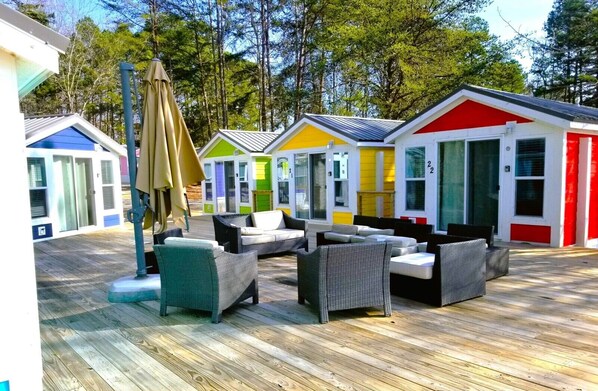 Colorful mobile homes line a serene natural setting, each with large windows and doors, a wooden deck, and comfortable outdoor furniture.