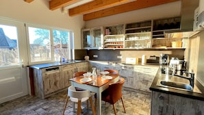 Une cuisine moderne avec des poutres en bois, un coin repas avec une table et des chaises, et une vue sur l’extérieur grâce à de grandes fenêtres.