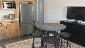 A kitchen with wooden cabinets, a stainless steel refrigerator, a microwave, a ceiling fan, a dining table with four chairs, and a flat-screen TV.