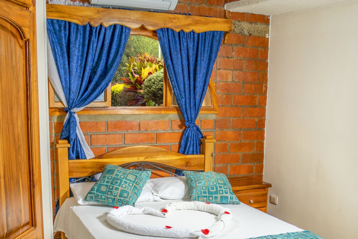 A bedroom with a brick wall, a wooden bed frame, a window with blue curtains, and an air conditioning unit above the bed.