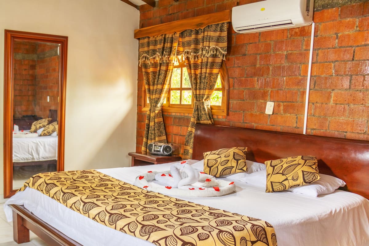 A hotel room with a brick wall, a bed with patterned bedding, a wooden headboard, and a window with curtains.
