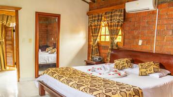 A hotel room with a brick wall, a bed with patterned bedding, a wooden headboard, and a window with curtains.