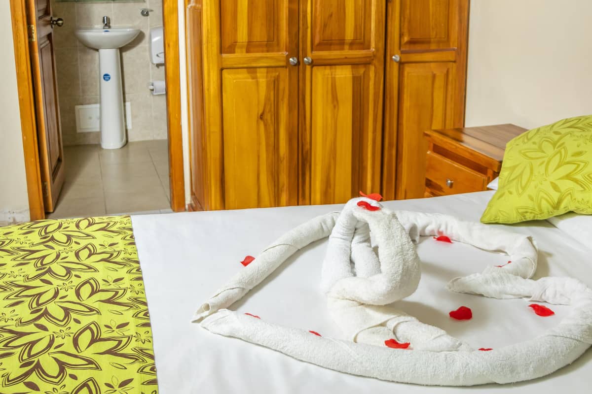 A hotel room with a wooden wardrobe, a bed with a white and yellow patterned bedspread, a green pillow, and a towel with red flowers.
