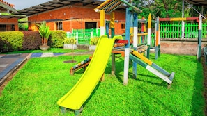A playground with a yellow slide and a climbing structure, surrounded by a brick building and a green fence. - hostal san jorge (Pereira)