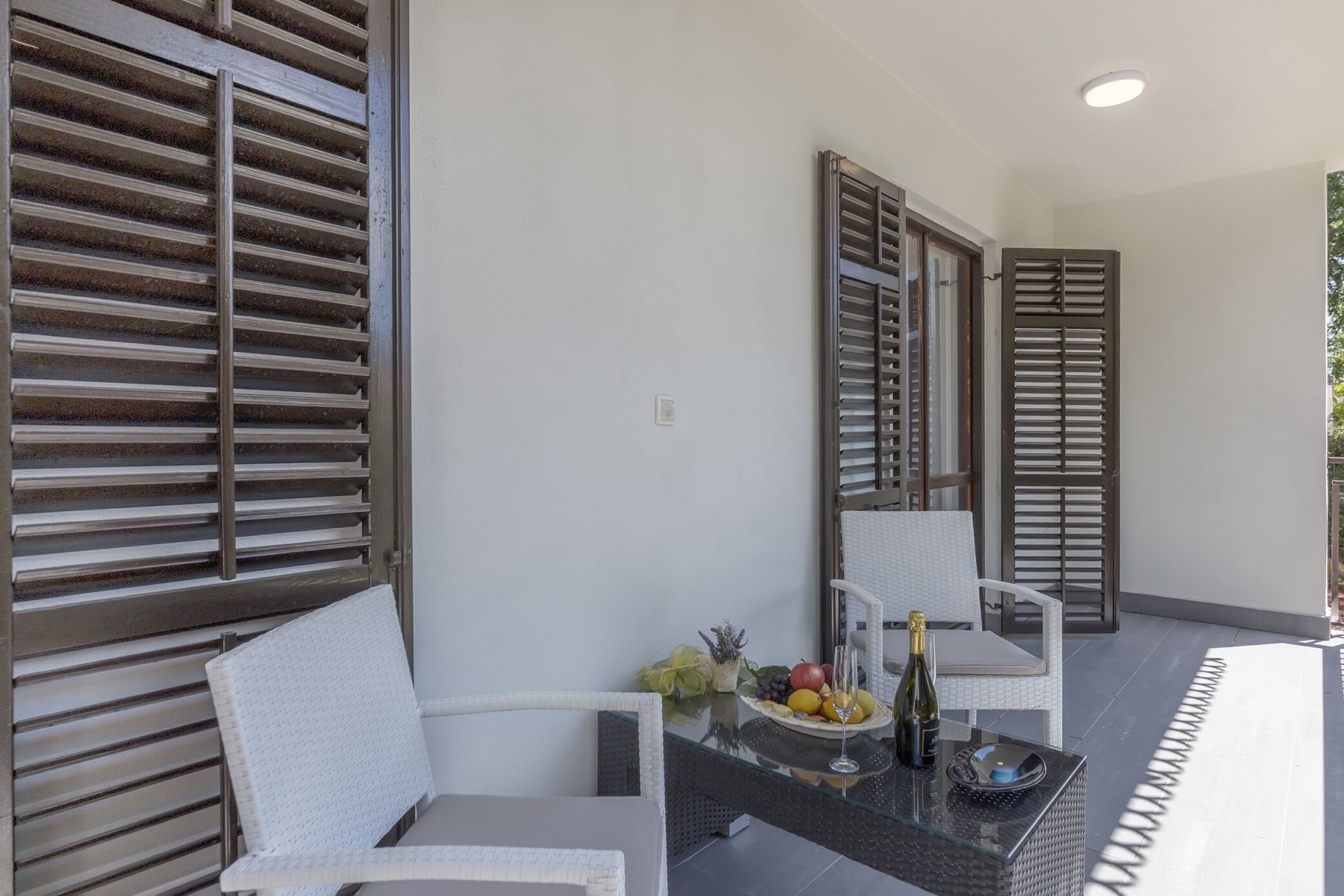 A balcony with white wicker furniture, a glass table with fruit and a bottle, and wooden shutters.