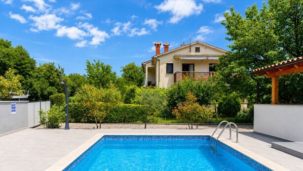 Une piscine bordée de carreaux, une maison avec un balcon et une végétation luxuriante tout autour.