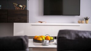 Un salon moderne avec un téléviseur à écran plat fixé au mur, une table basse sur laquelle se trouve un panier de fruits et une tasse, ainsi que des fauteuils en cuir foncé.