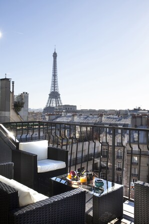 Una terraza en la azotea con mobiliario de mimbre, una mesa con una bandeja de frutas y una tetera, y una vista de la Torre Eiffel al fondo.