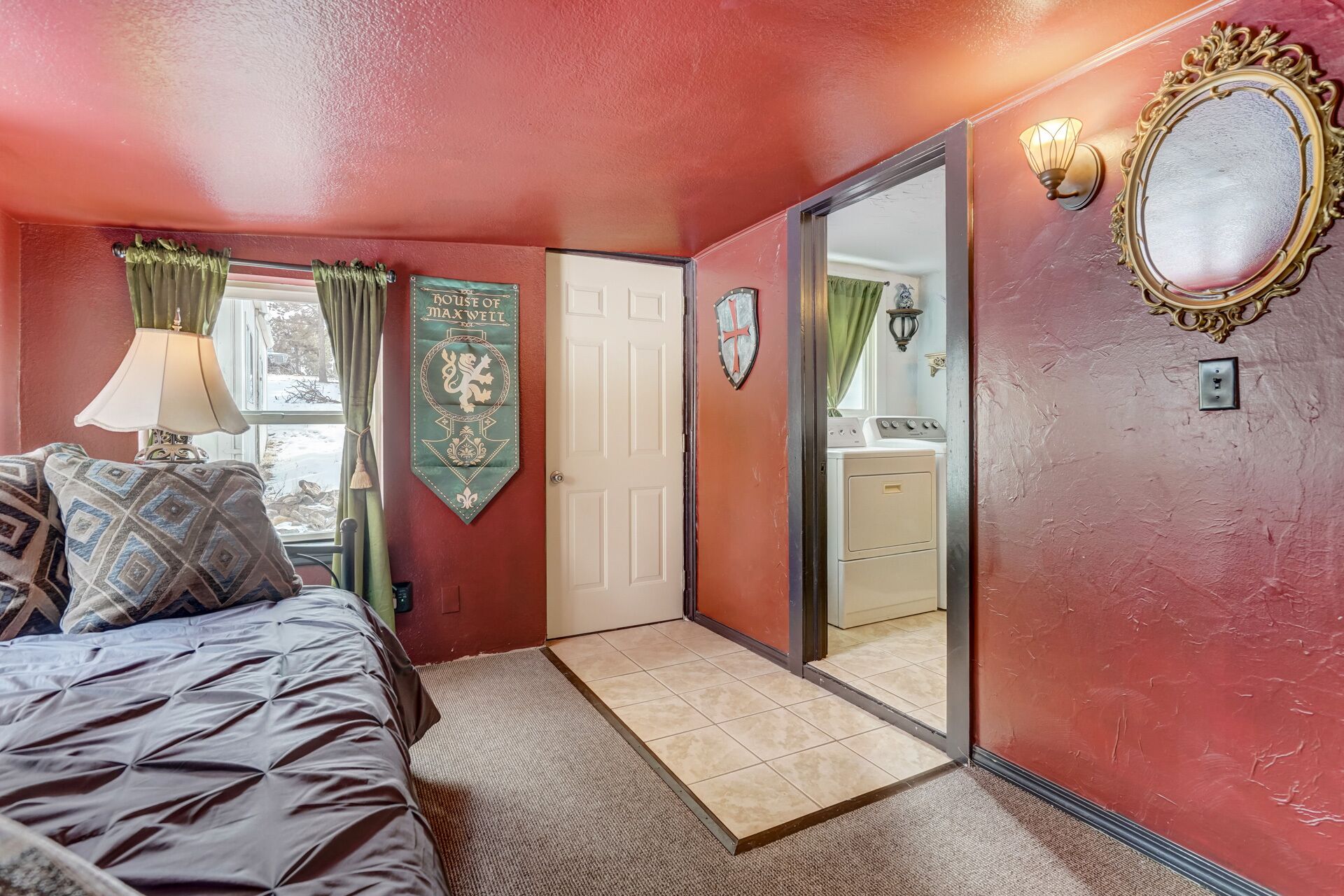 A bedroom with a bed, a window with curtains, a coat of arms, a mirror, and a door leading to a bathroom.