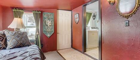Un dormitorio con cama, ventana con cortinas, escudo, espejo y puerta que da a un baño.