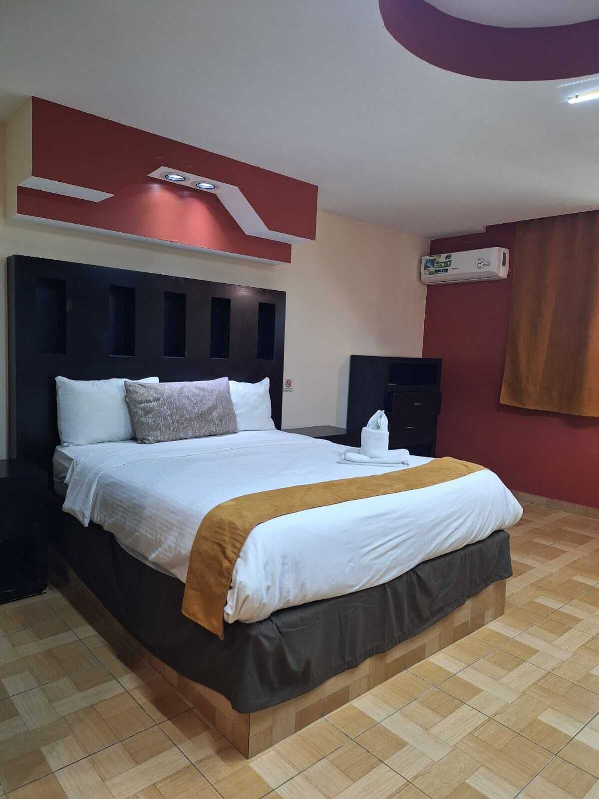 Quarto de hotel com uma cama grande, cabeceira escura, ar condicionado fixado na parede e piso de madeira.