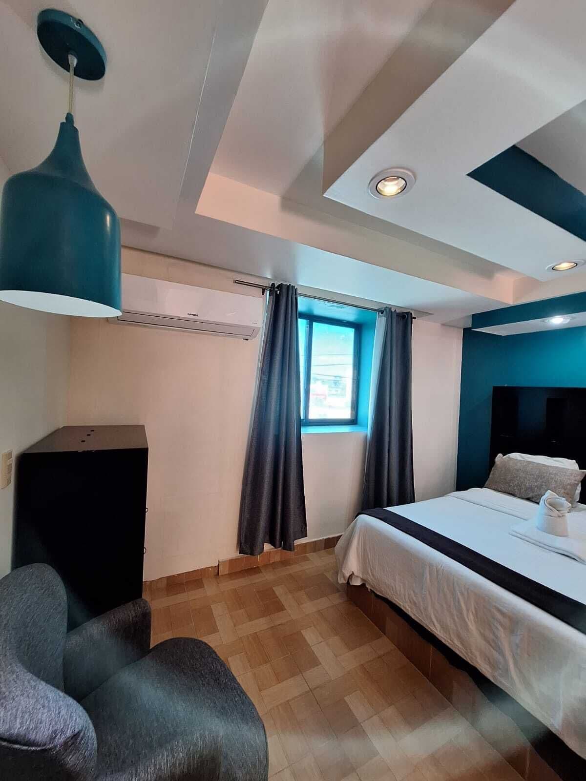Una habitación de hotel moderna con cama, escritorio y silla. Cuenta con ventana con cortinas, una lámpara en el techo y aire acondicionado de pared.
