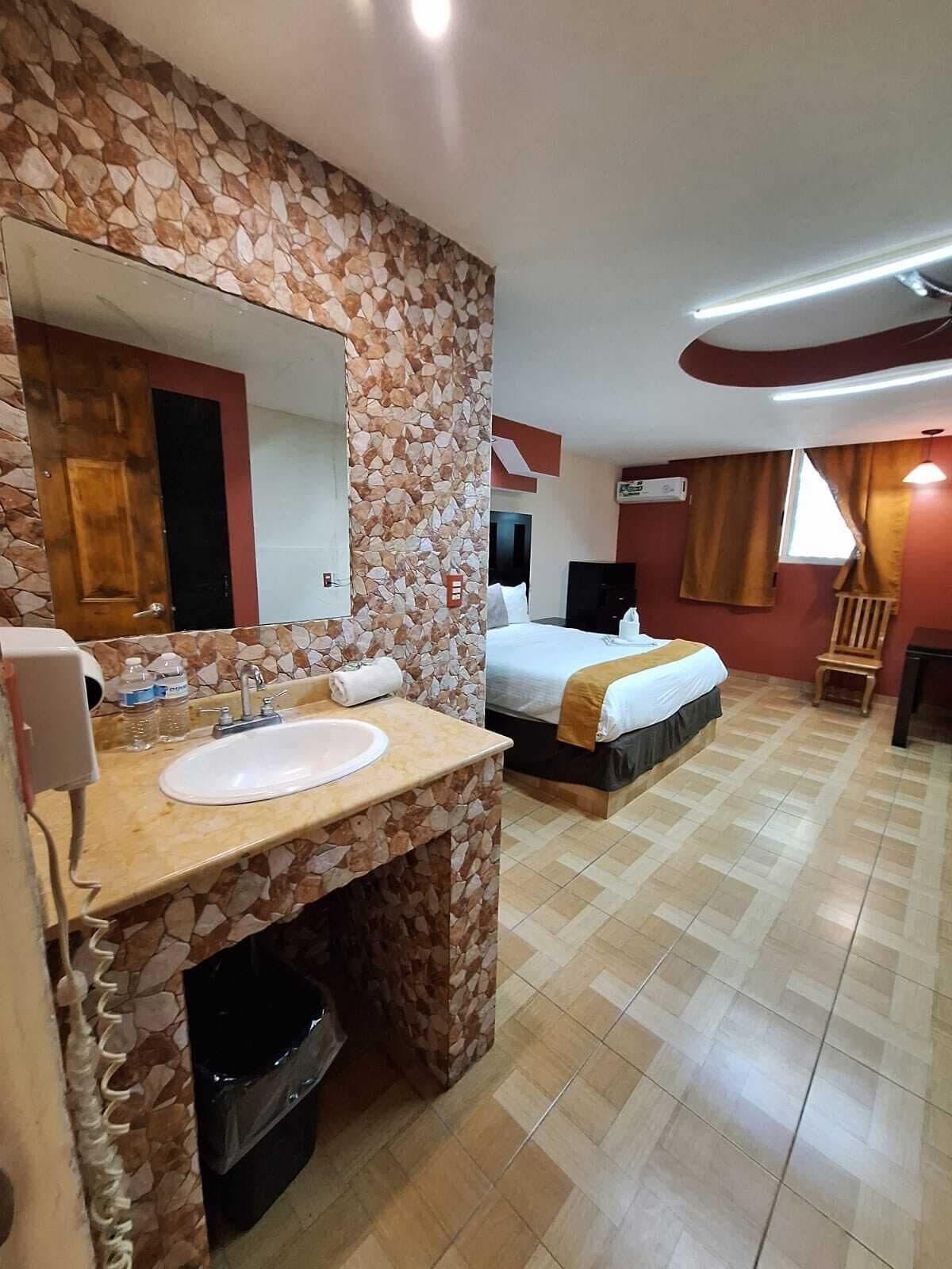 A hotel room with a stone-patterned wall, a large mirror, a sink, a bed, a TV, and a window with curtains.