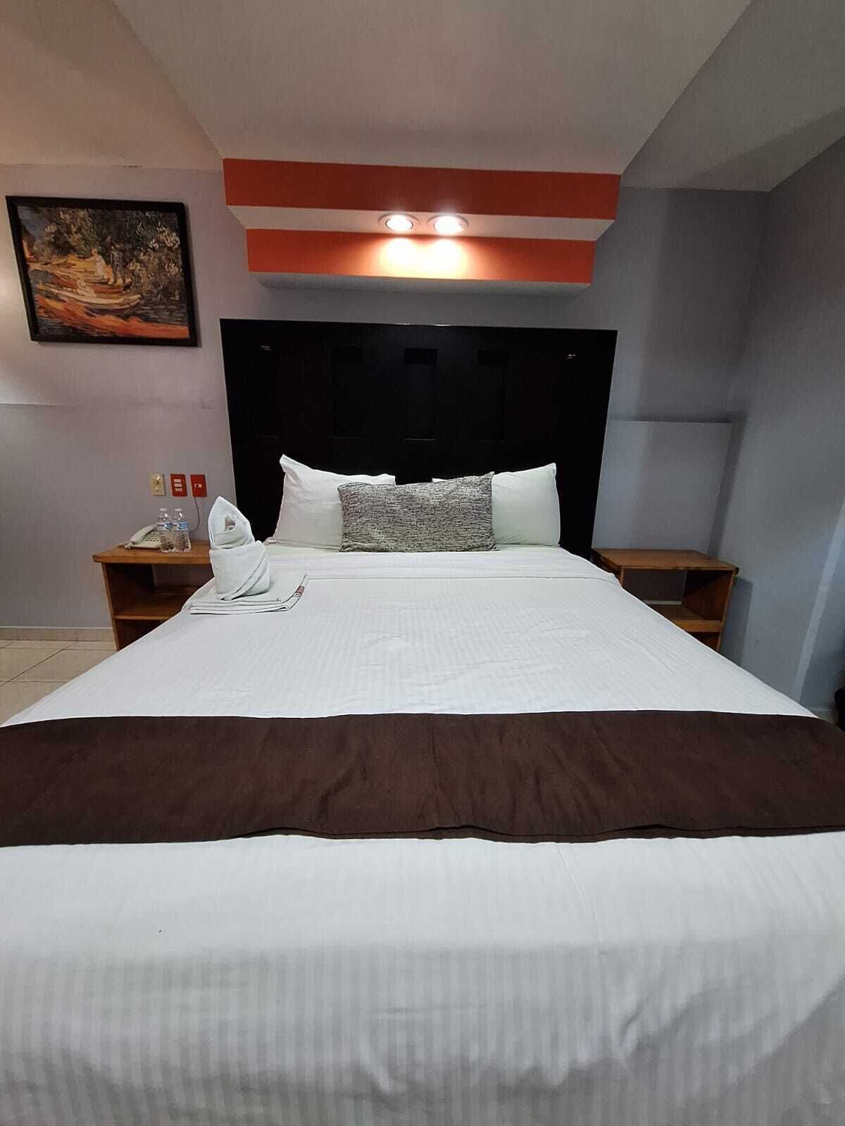 Une chambre d’hôtel avec un grand lit, une tête de lit dotée d’un éclairage intégré, un tableau au mur et une petite table sur laquelle se trouvent un téléphone et des bouteilles d’eau.