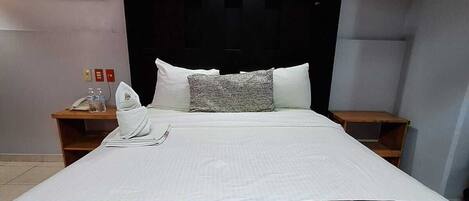 Habitación de hotel con una cama grande, cabecera con iluminación integrada, un cuadro en la pared y una mesita con teléfono y botellas de agua.