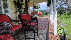 Una terraza de madera con mobiliario de mimbre y un tapete, una casa amarilla con porche, y un jardín al fondo.