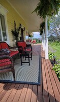 A wooden deck with wicker furniture and a rug, a yellow house with a porch, and a garden in the background.