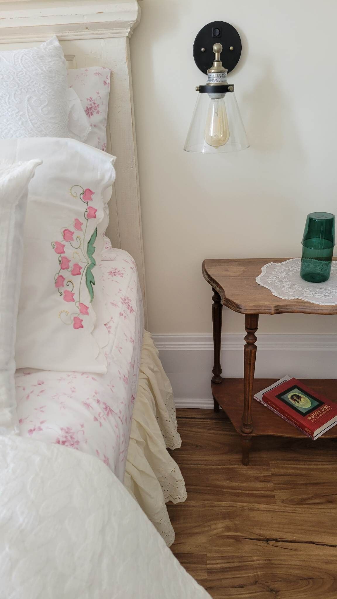 Una cama bien hecha con ropa de cama floral, una mesita de noche con un libro y una lámpara fijada a la pared.