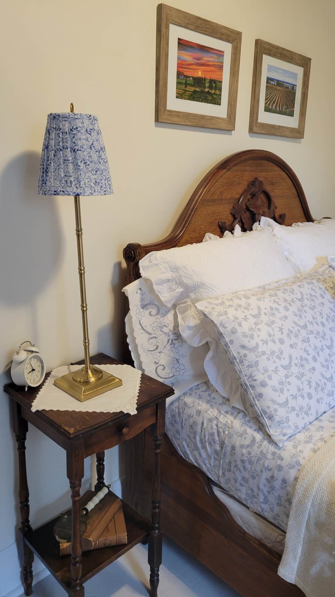 Quarto com cama de madeira, mesa de cabeceira com abajur, relógio e duas fotos emolduradas na parede.