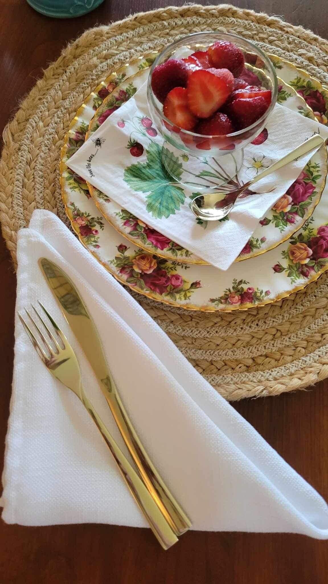 A table setting with a woven placemat, a floral-patterned plate, a glass bowl of strawberries, a folded napkin, and a gold fork and spoon.