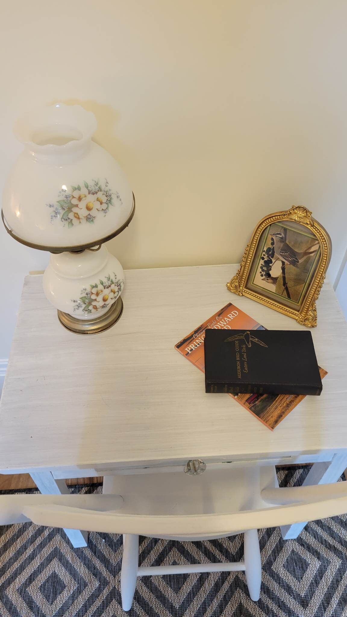 Une petite table blanche avec une lampe, un tableau encadré et un livre.