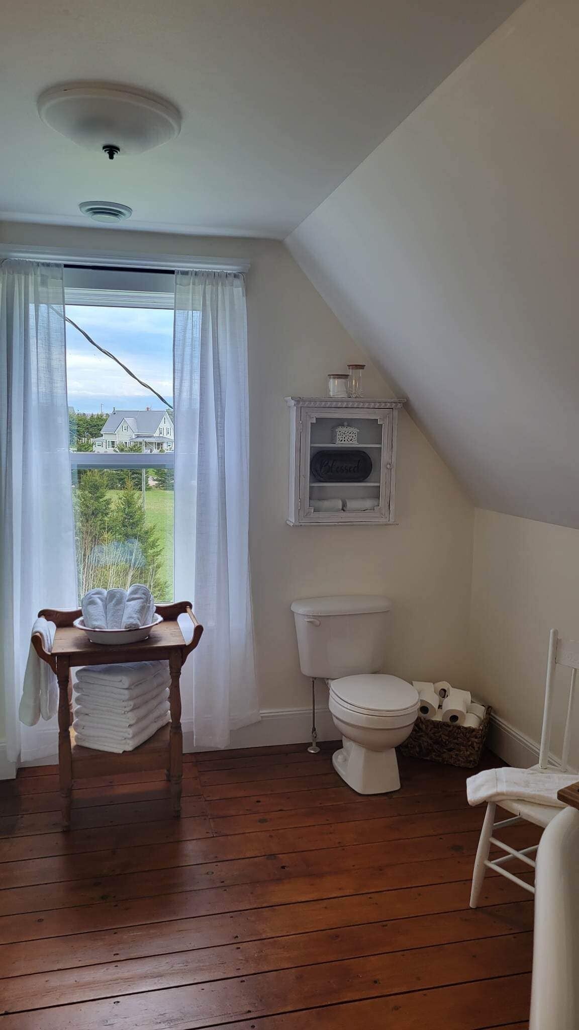 A bathroom with a toilet, a small table with towels, a window with curtains, and a view of a house and trees outside.