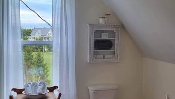 A bathroom with a toilet, a small table with towels, a window with curtains, and a view of a house and trees outside.