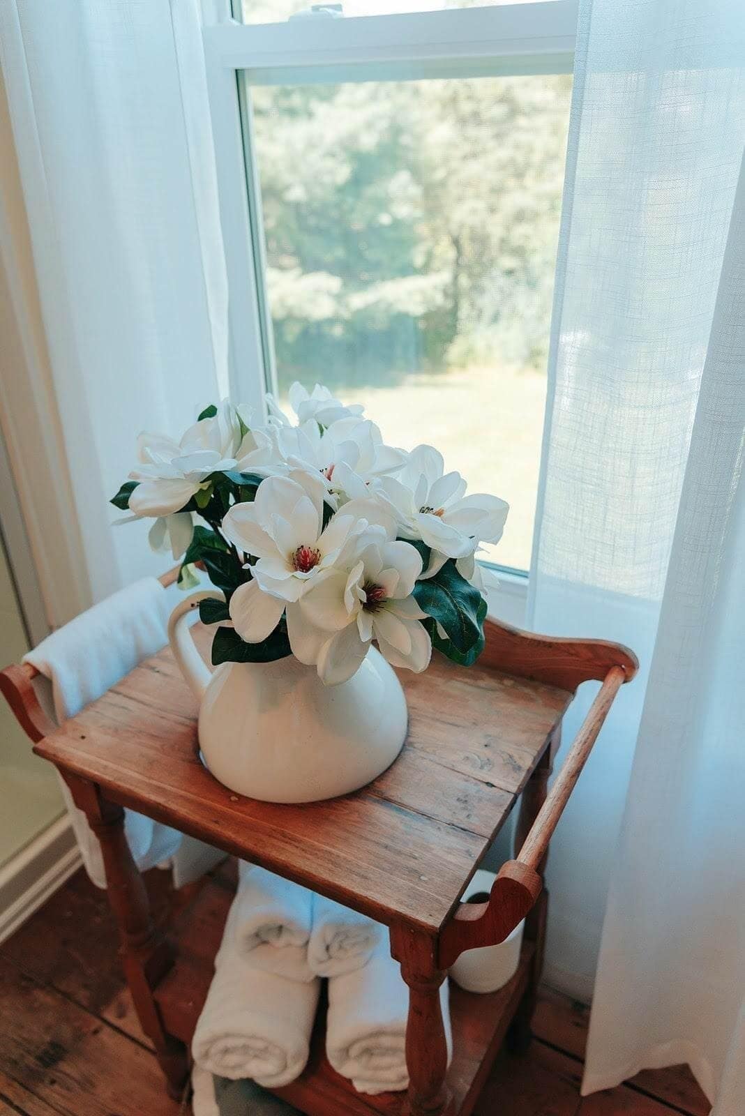 A white vase with white flowers on a wooden table by a window.