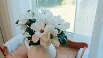 A white vase with white flowers on a wooden table by a window.